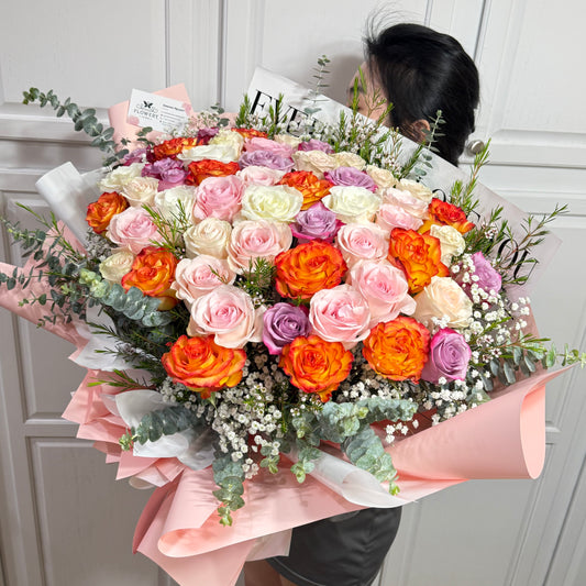 Vibrant bouquet of pink, orange, white, and purple roses wrapped in pink paper.
