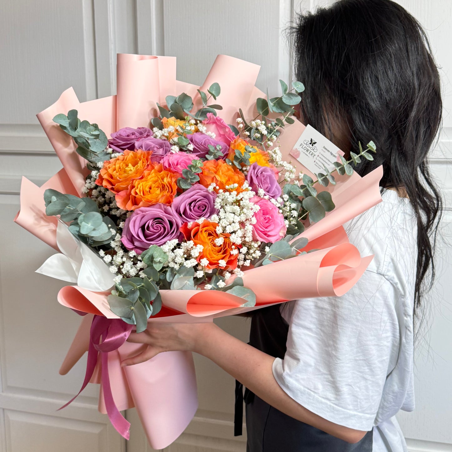 Vibrant bouquet of purple, orange, and pink roses wrapped in light pink paper.
