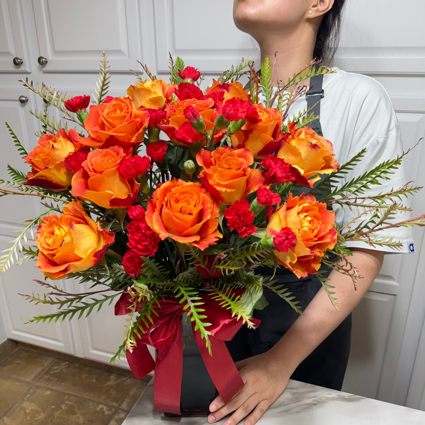 Vibrant orange roses and red carnations in a dark vase with a red ribbon.