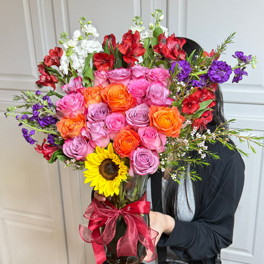 Vibrant bouquet of roses, sunflower, and mixed flowers in a clear vase.