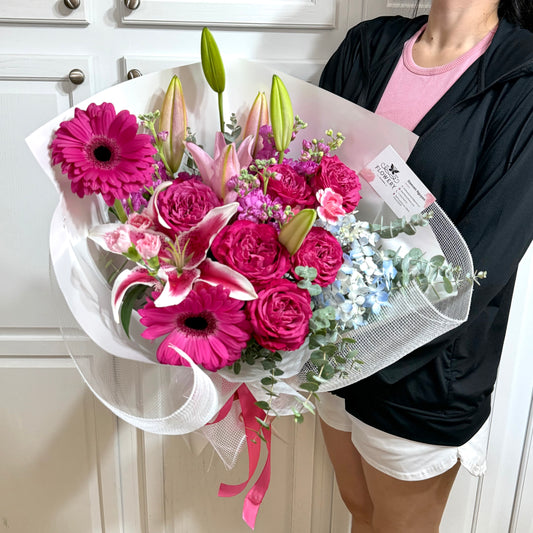 Vibrant pink lilies, roses, and daisies bouquet wrapped in white mesh.