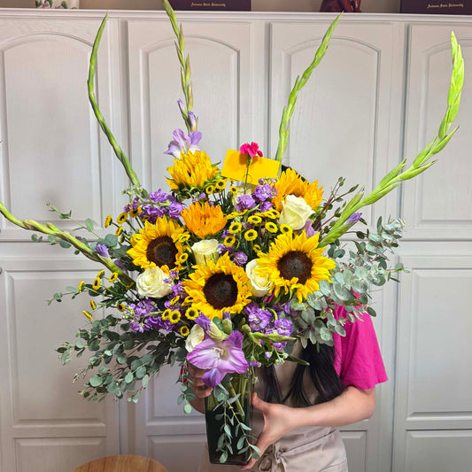 Large vibrant bouquet with yellow sunflowers, white roses, purple flowers, and green foliage.