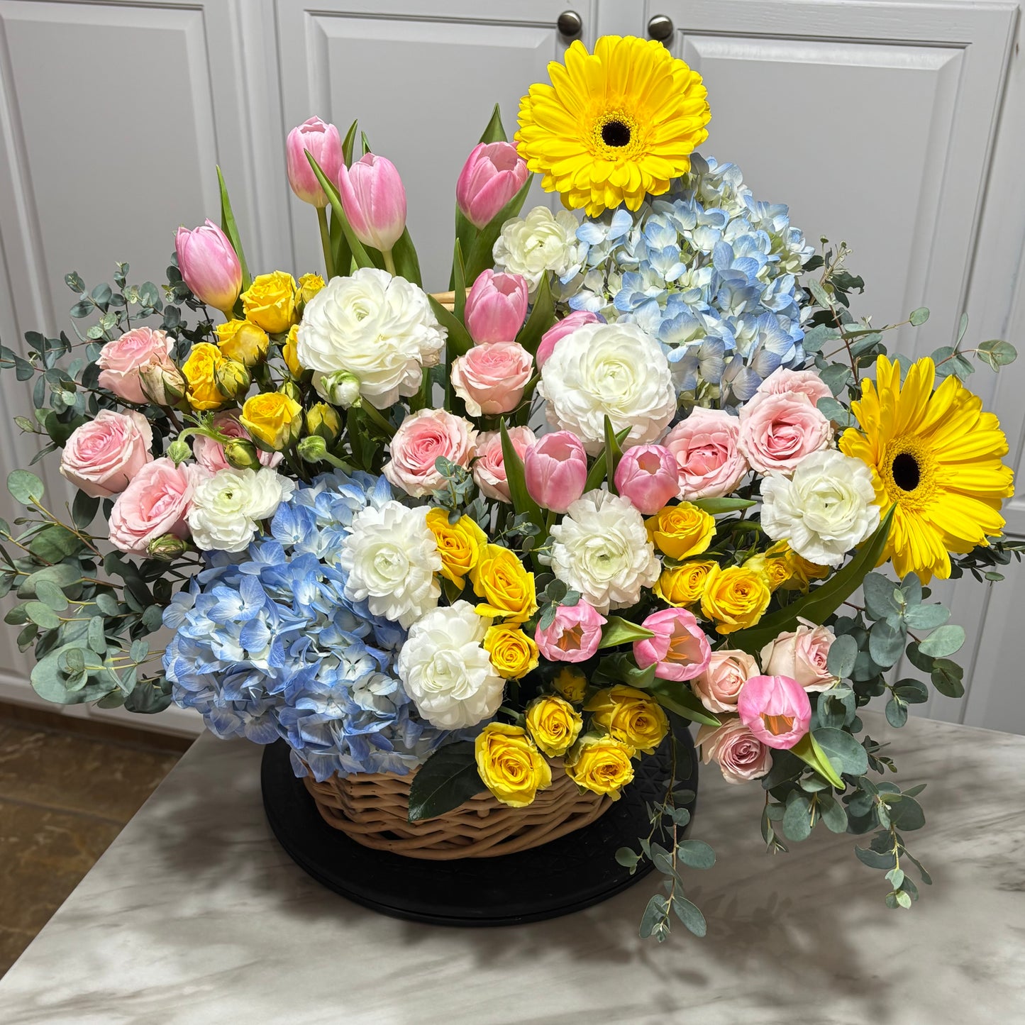 Vibrant spring flower arrangement in a wicker basket with pink, yellow, and blue blooms.
