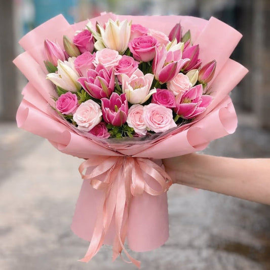 A hand holds a pink bouquet of roses, tulips, and white lilies.