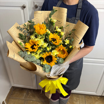 Vibrant yellow sunflowers and small flowers in a newspaper-wrapped bouquet with a yellow ribbon.