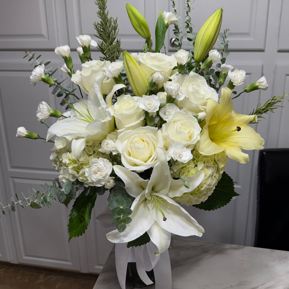 A full white and yellow floral arrangement with roses, lilies, and green leaves.