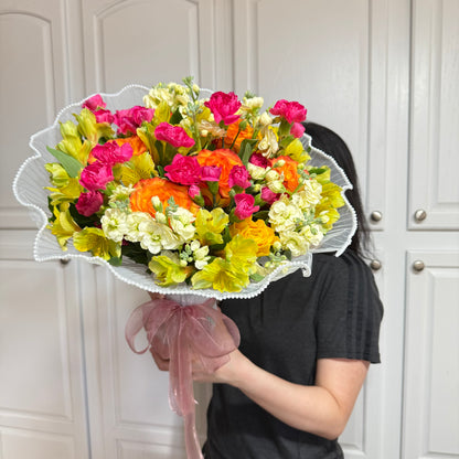 Vibrant mixed flower bouquet with pink, orange, yellow, and white blooms in white netting.