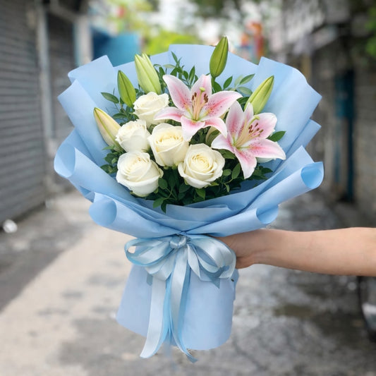 A hand holds a bouquet of white roses and pink lilies wrapped in light blue paper.