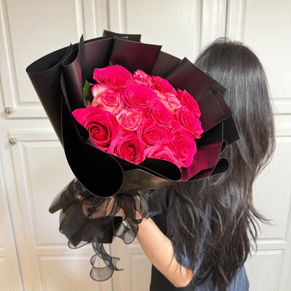 Vibrant pink roses in a large bouquet wrapped in elegant black paper and ribbon.