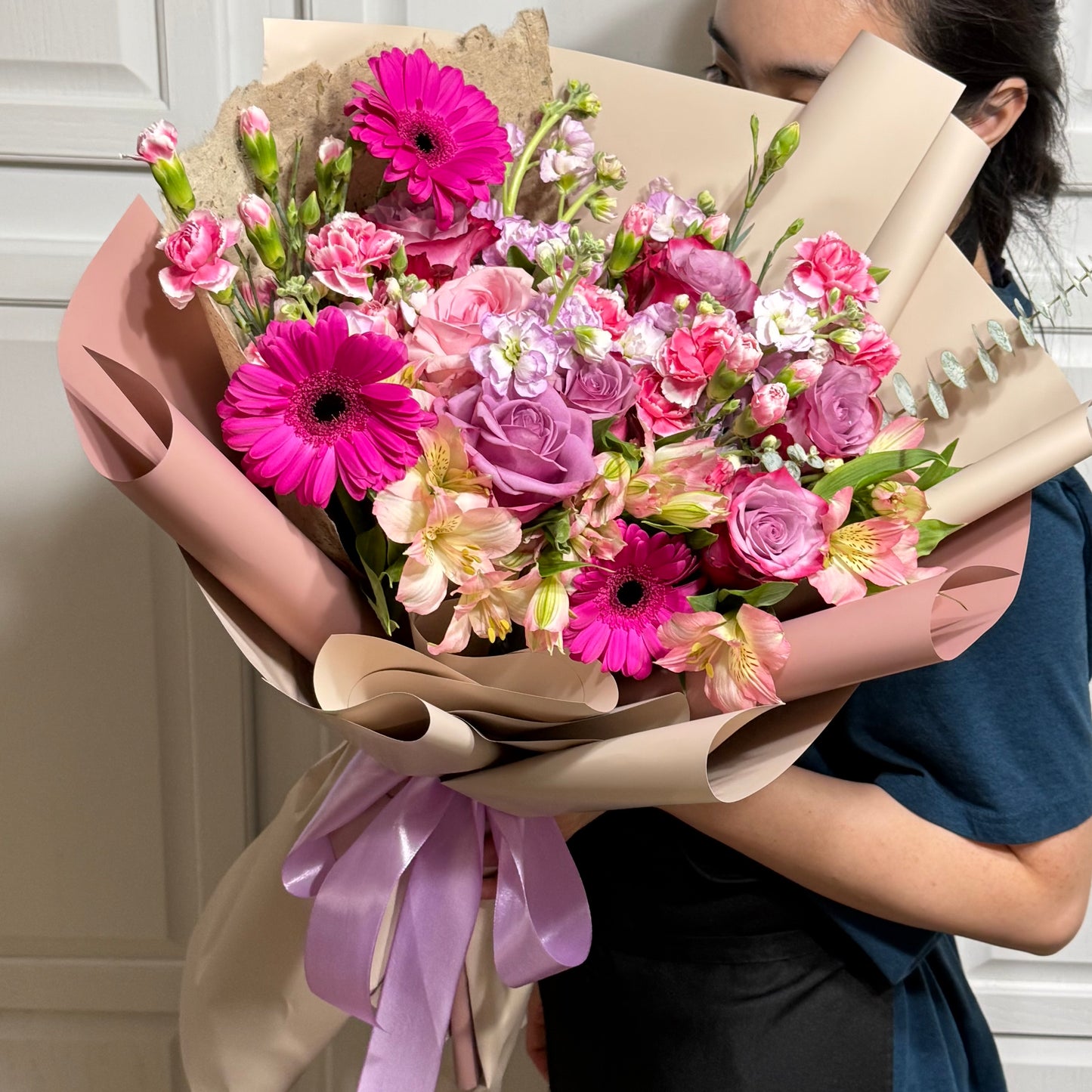 Large bouquet of pink gerberas, purple roses, and mixed flowers in pink and beige wrap.