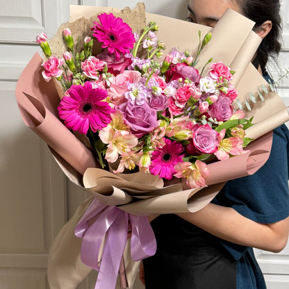 Large bouquet of pink gerberas, purple roses, and mixed flowers in pink and beige wrap.