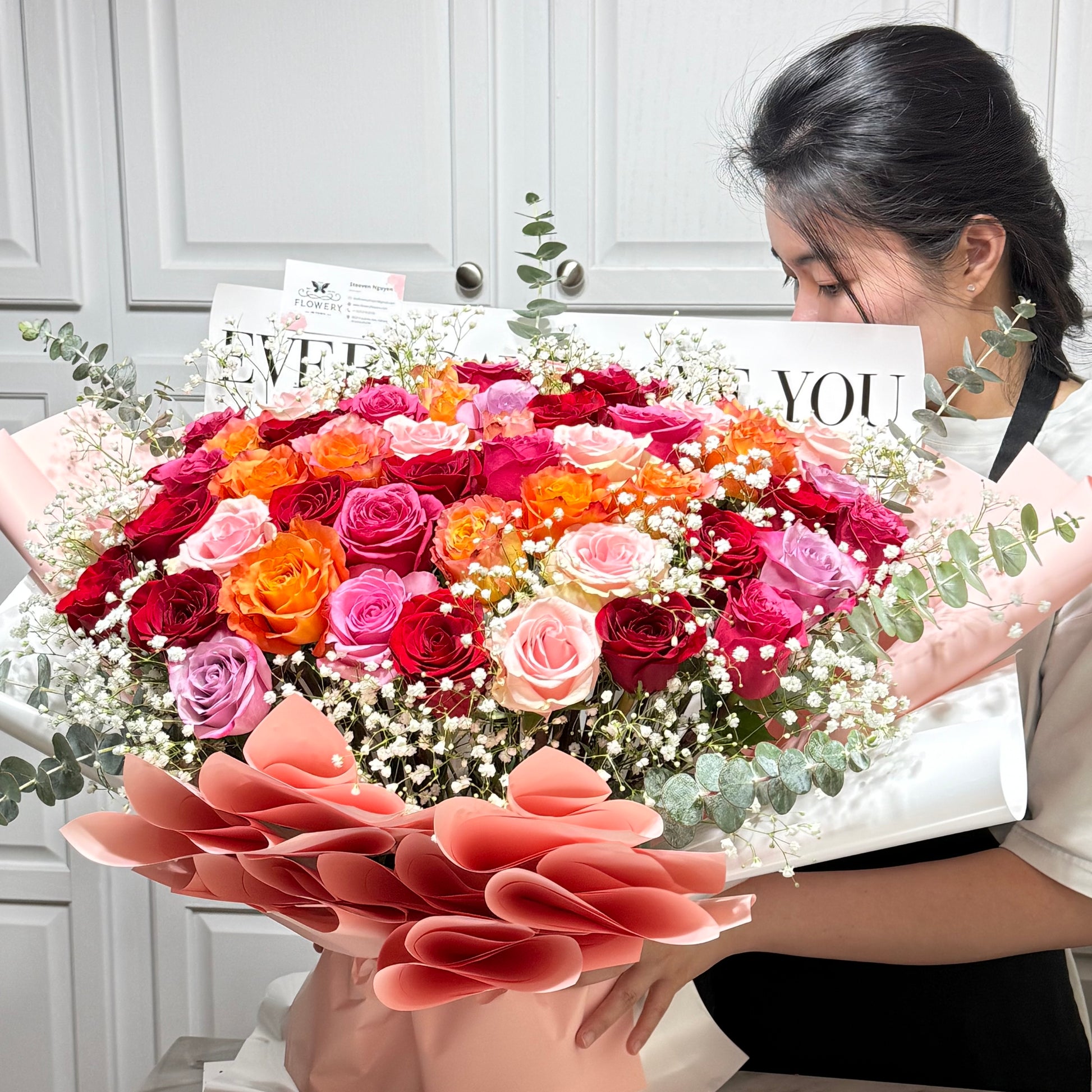 Large bouquet of red, orange, and pink roses with white filler held by a woman.