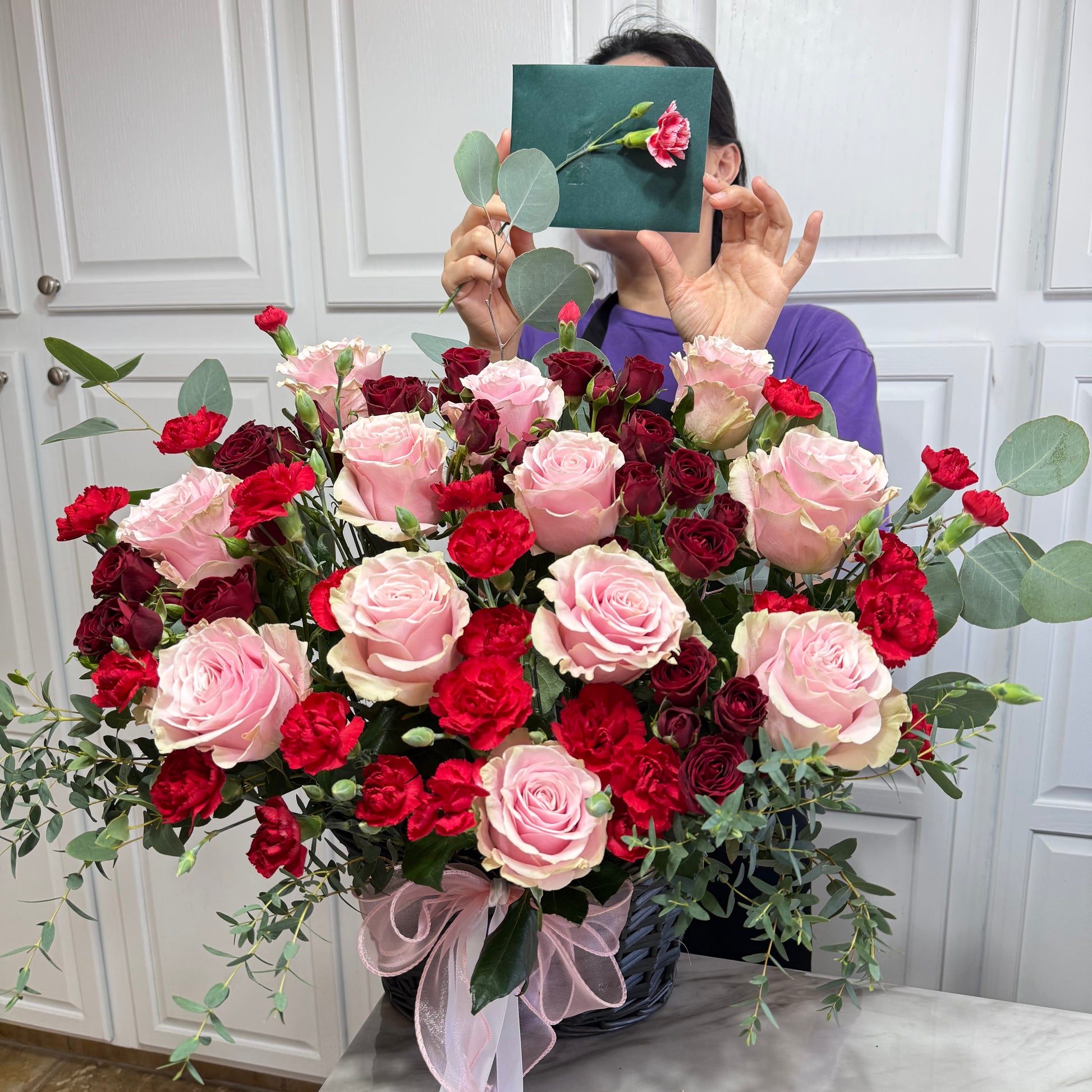 Large basket of pink roses, red carnations, and eucalyptus with a pink bow.