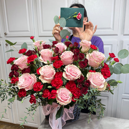 Large basket of pink roses, red carnations, and eucalyptus with a pink bow.