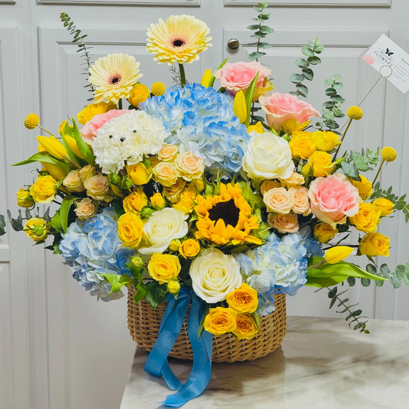 Colorful spring flowers, including a dog-shaped one, in a woven basket with blue ribbon.