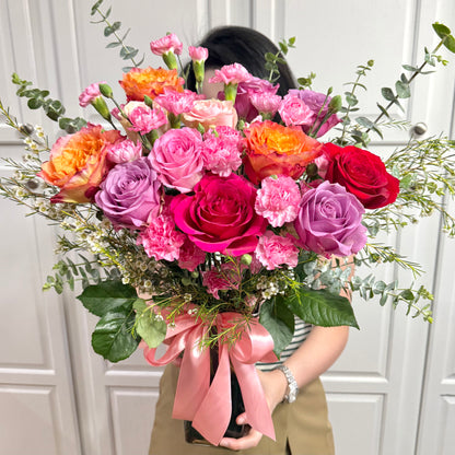 Vibrant bouquet of pink, purple, orange, and red roses and carnations with green foliage.