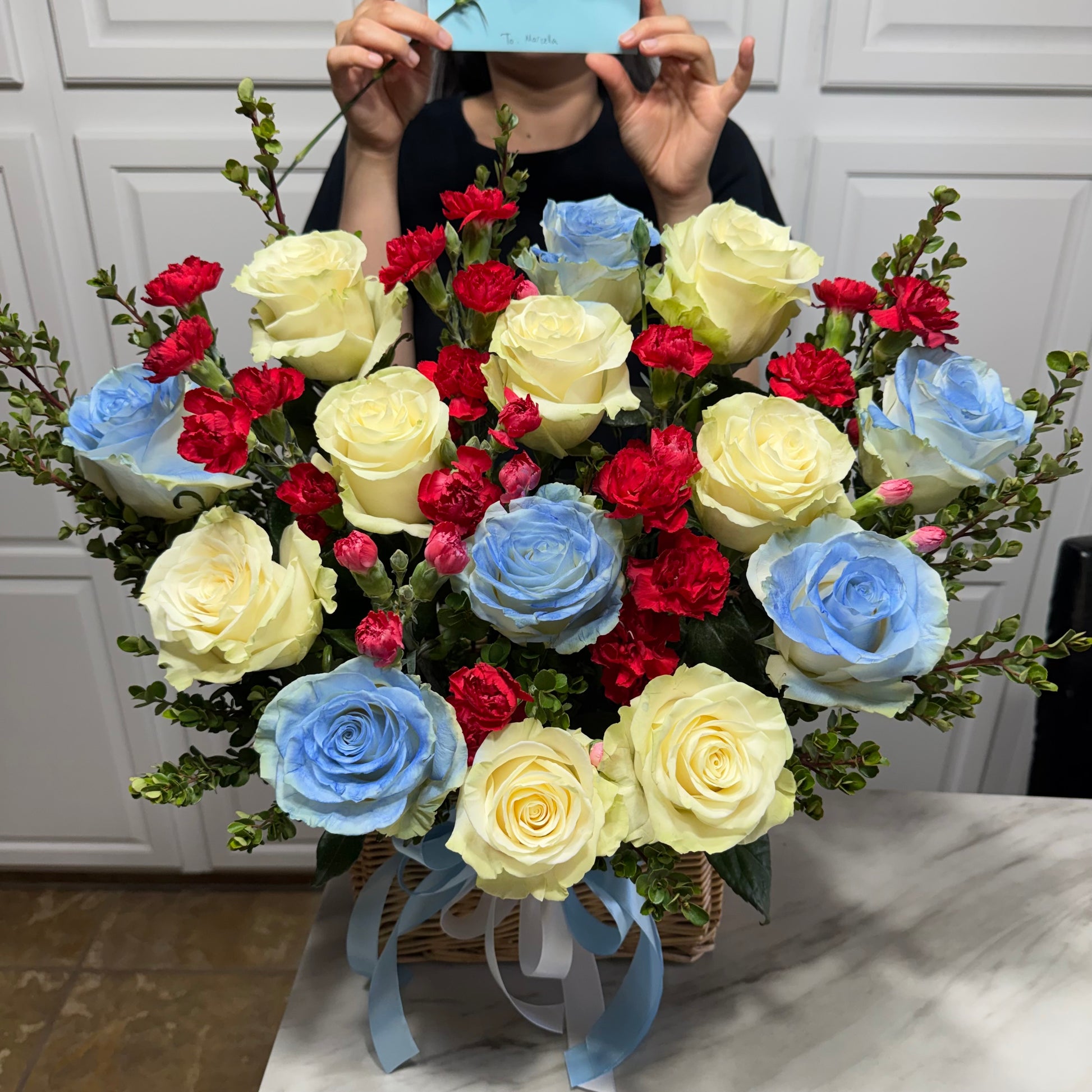 Vibrant bouquet featuring yellow and blue roses, red carnations, and greenery in a basket.