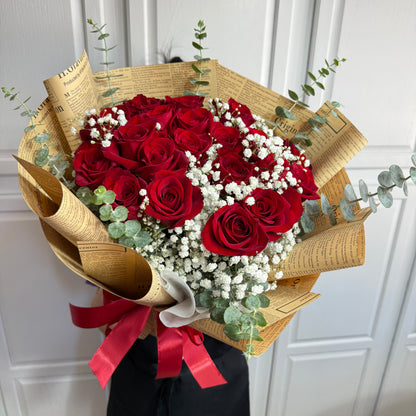 Vibrant red roses and white baby's breath in a newspaper-wrapped bouquet with a red ribbon.