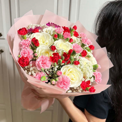 A person holds a vibrant bouquet of white roses, pink and red carnations, wrapped in pink.