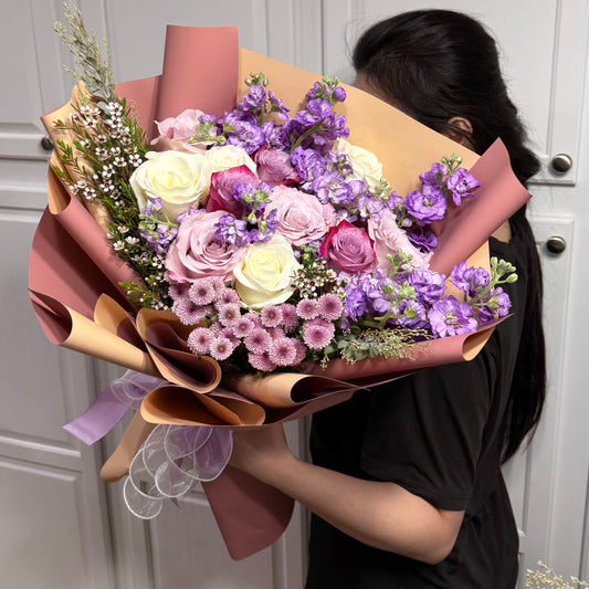 Vibrant bouquet of white, pink, purple roses and stock, wrapped in elegant two-toned paper.
