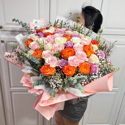 Large bouquet of colorful roses, baby's breath, and eucalyptus wrapped in pink paper.