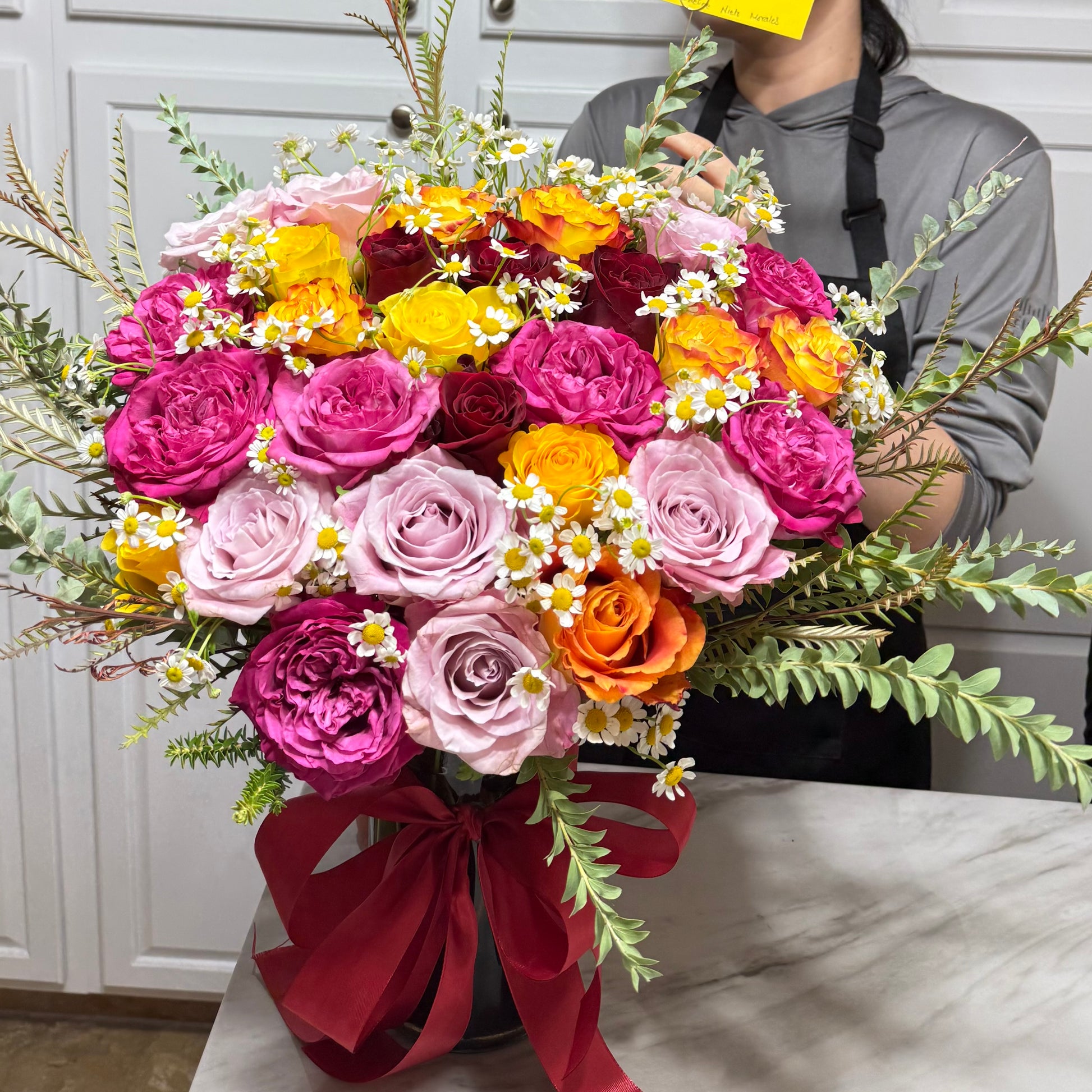 Large colorful rose bouquet with daisies and green foliage in a red-ribboned vase.
