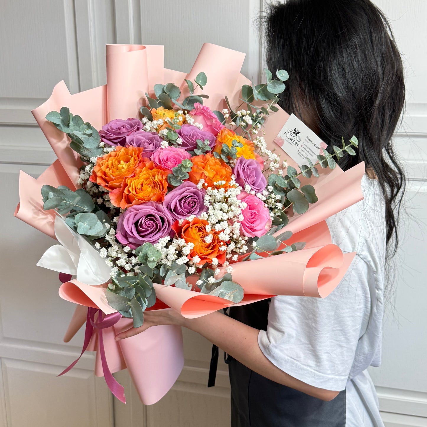 Vibrant bouquet of orange, purple, and pink roses wrapped in pink paper.