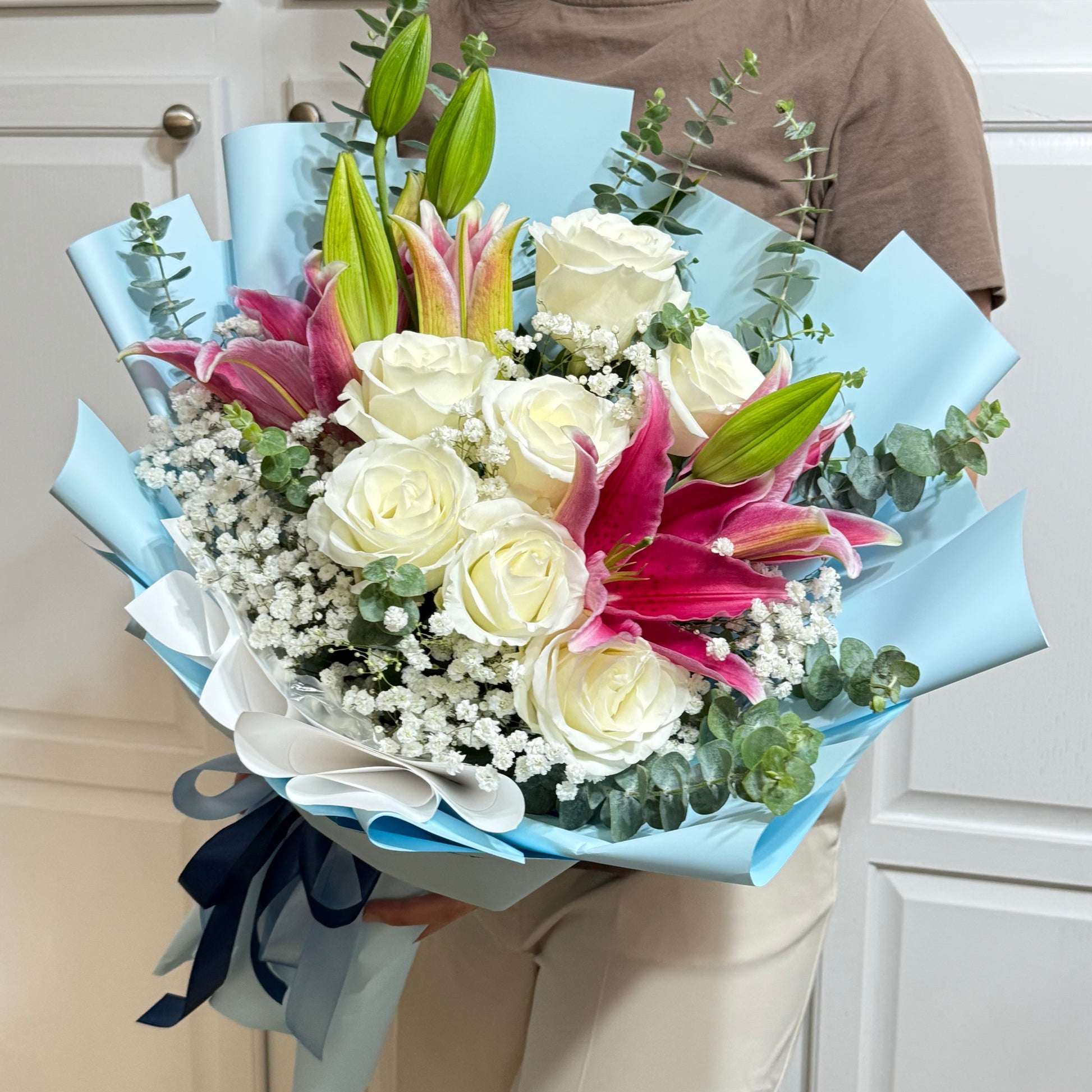 A bouquet of white roses, pink lilies, baby's breath, and eucalyptus wrapped in blue paper.