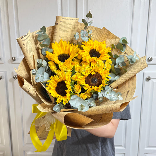 Vibrant sunflower and yellow rose bouquet wrapped in newspaper print with a yellow ribbon.