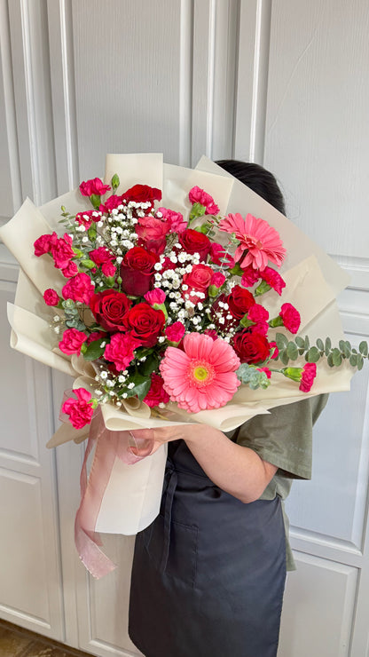 A person holds a large bouquet of red roses, pink carnations, and a gerbera daisy.