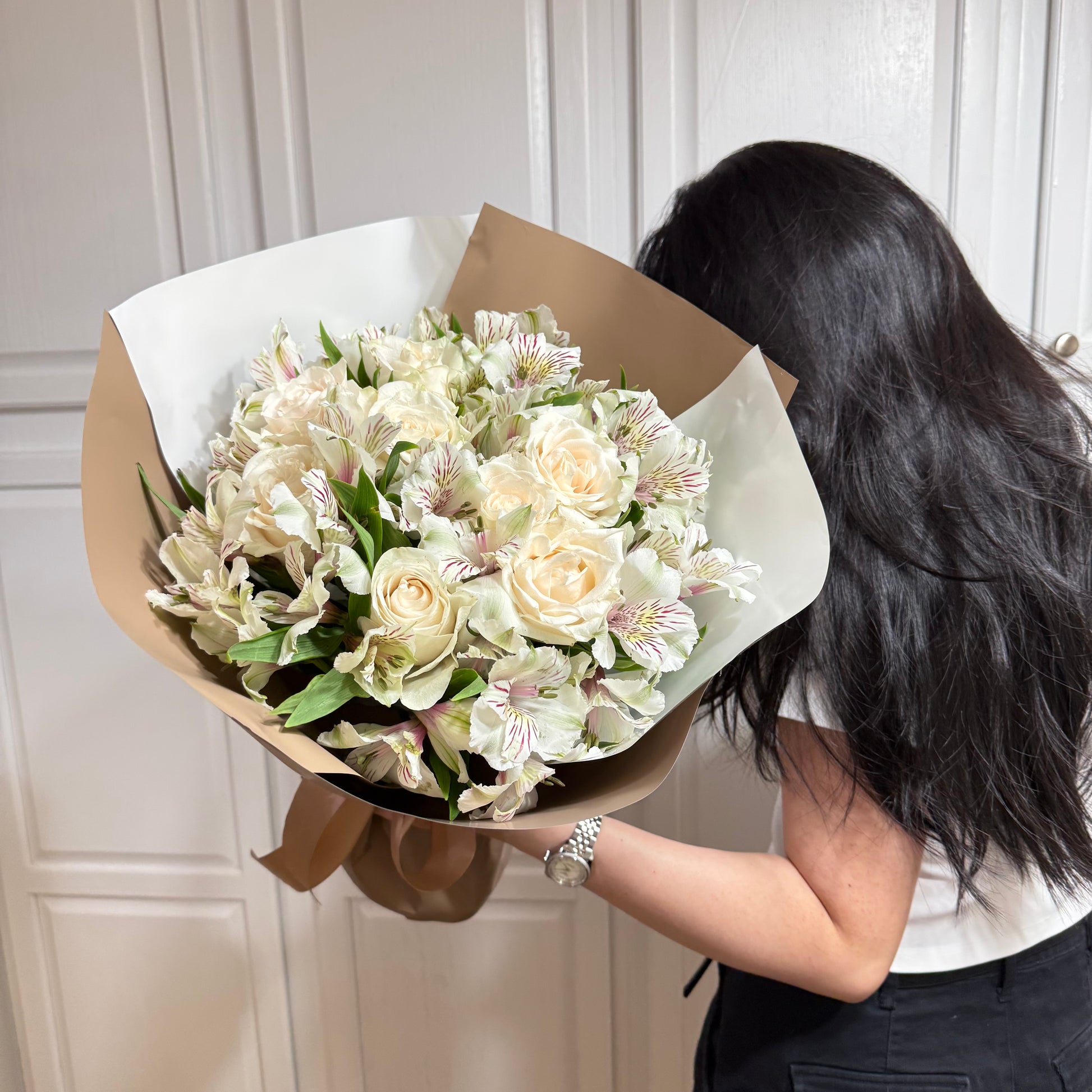 White roses and striped alstroemeria bouquet wrapped in brown paper, held by a person.