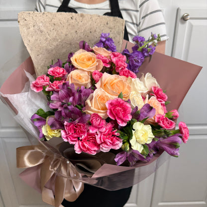 Vibrant bouquet of peach roses, pink carnations, and purple alstroemeria, wrapped in brown paper.