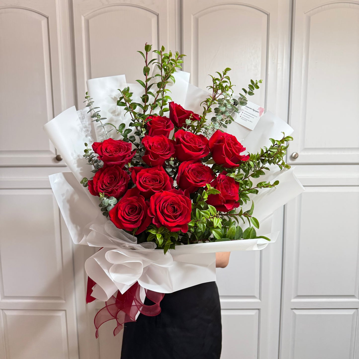 Vibrant red rose bouquet with green foliage, wrapped in white paper and red ribbon.
