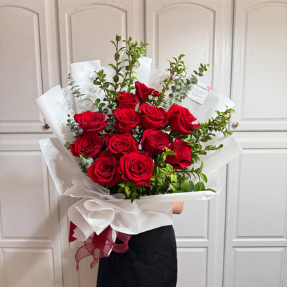 Vibrant red rose bouquet with green foliage, wrapped in white paper and red ribbon.