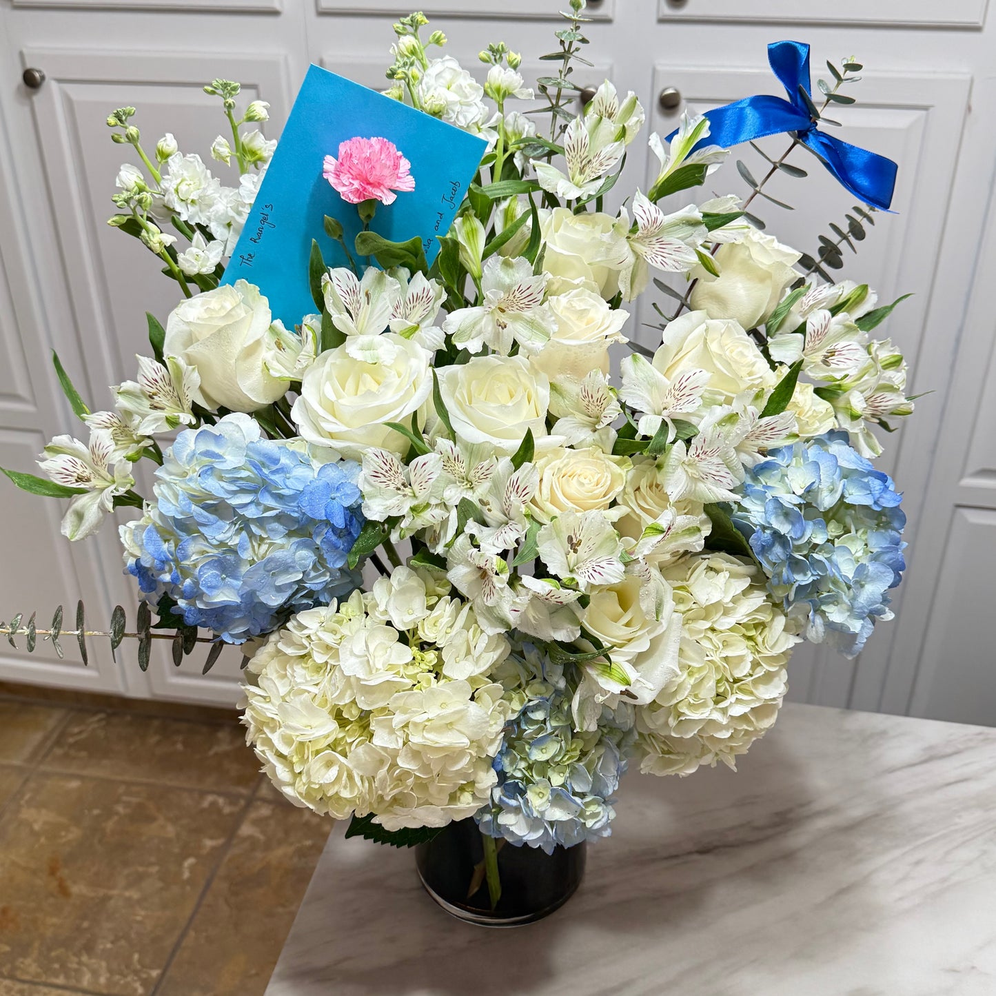 White and blue floral bouquet with roses, hydrangeas, and a blue ribbon.