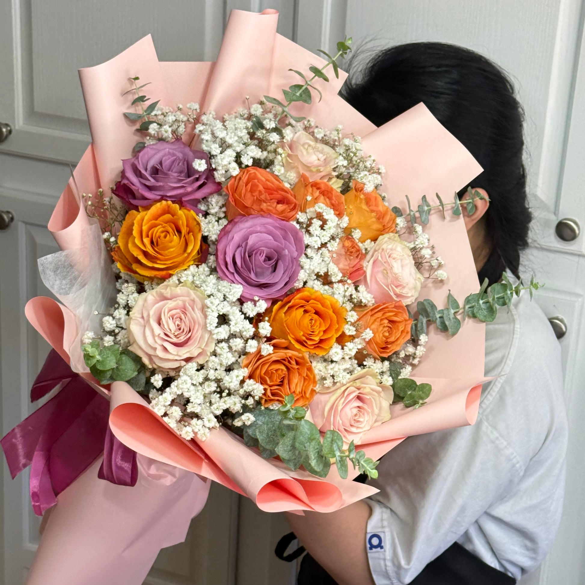 A vibrant bouquet of purple, orange, and pink roses with baby's breath, wrapped in pink paper.