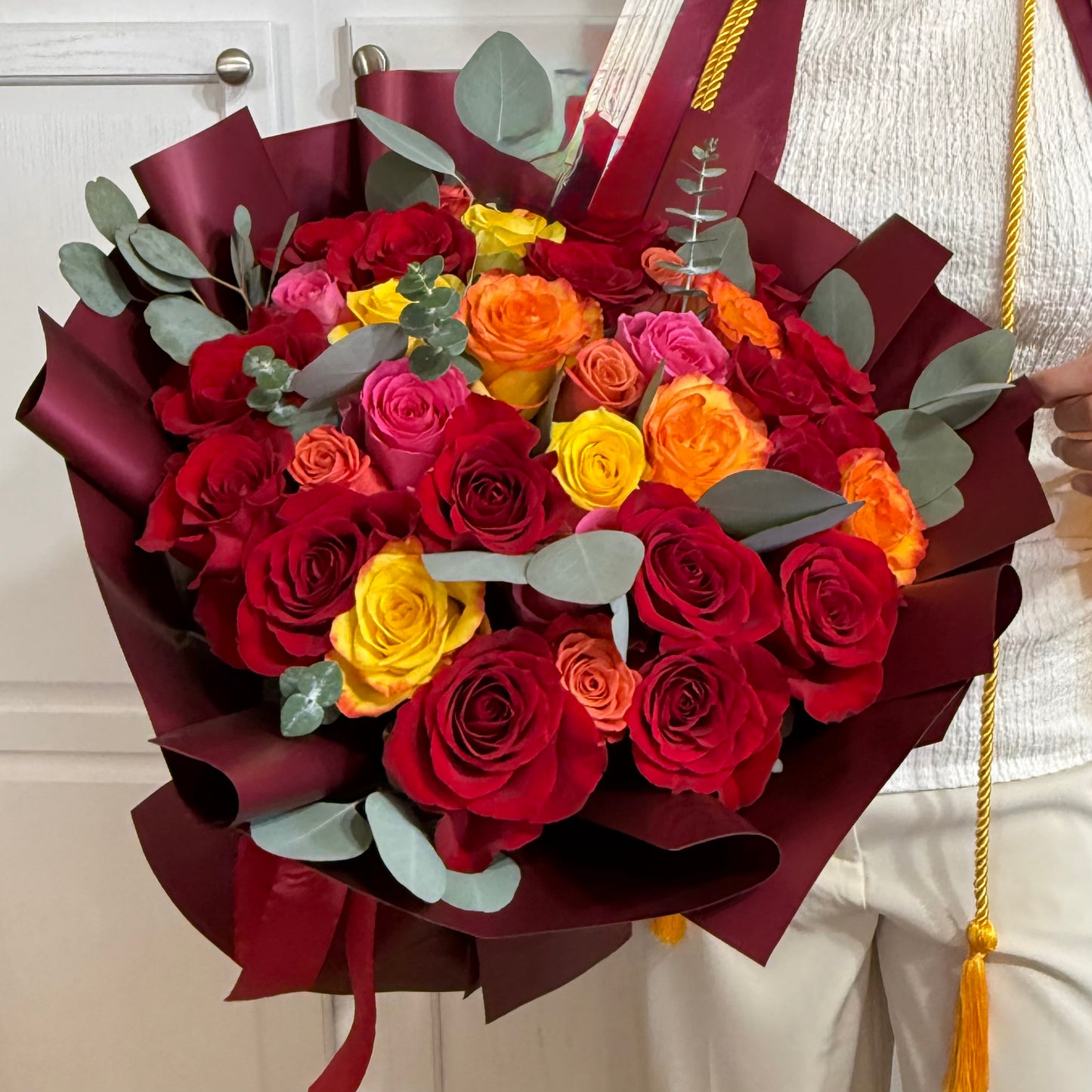Vibrant red, orange, and yellow rose bouquet wrapped in burgundy paper.