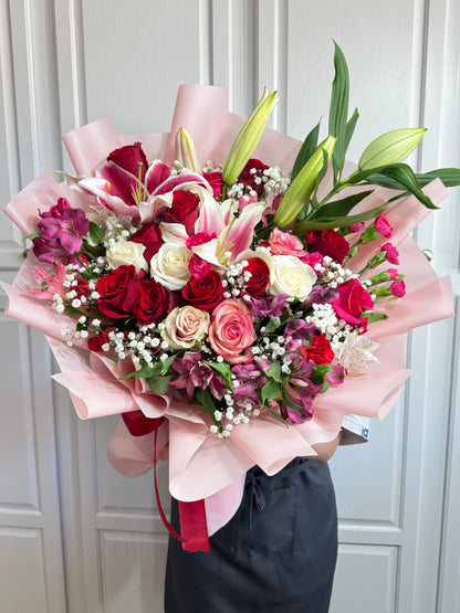 Large bouquet of red, white, and pink roses, lilies, and filler flowers wrapped in pink.