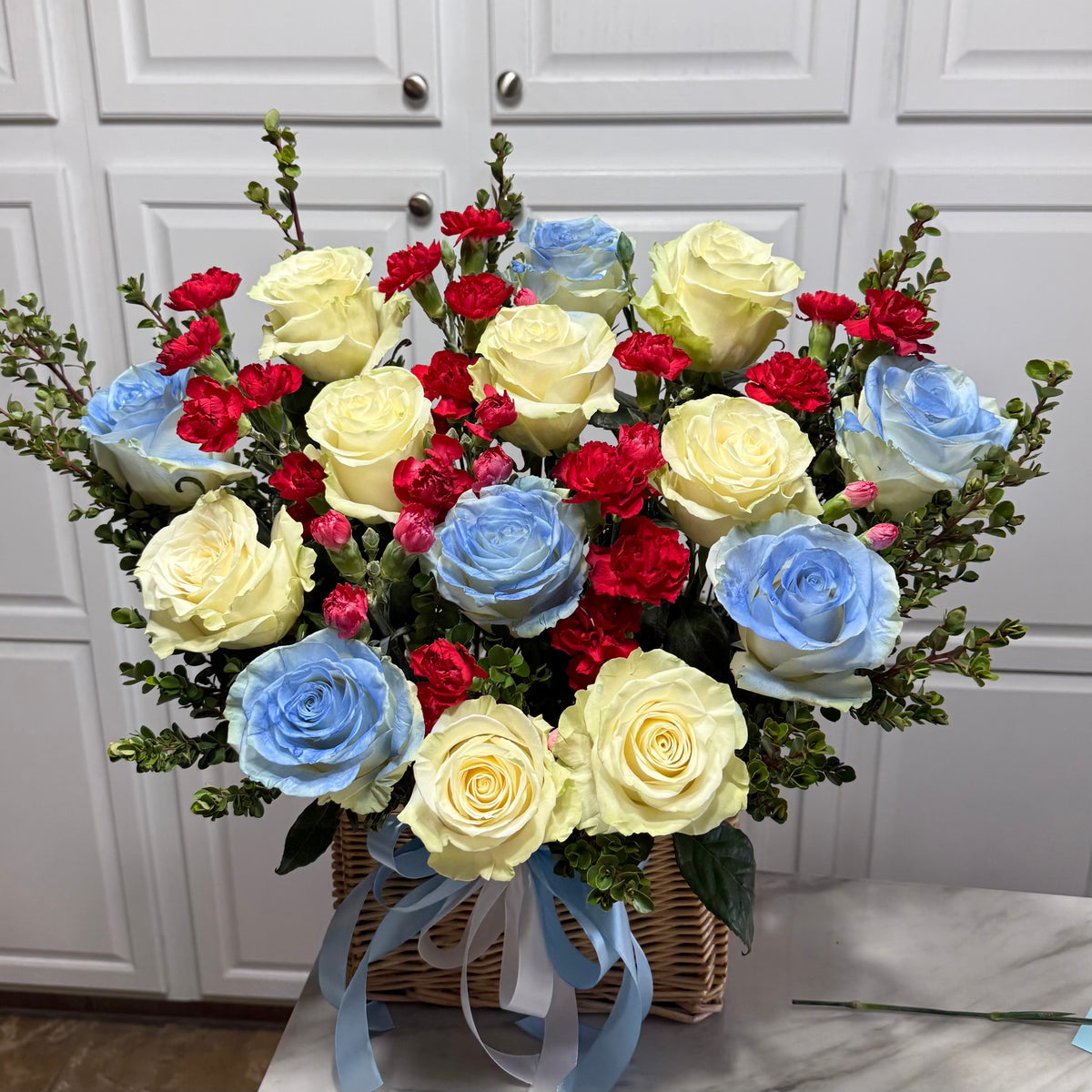 A large basket of yellow and blue roses with red carnations and green foliage.