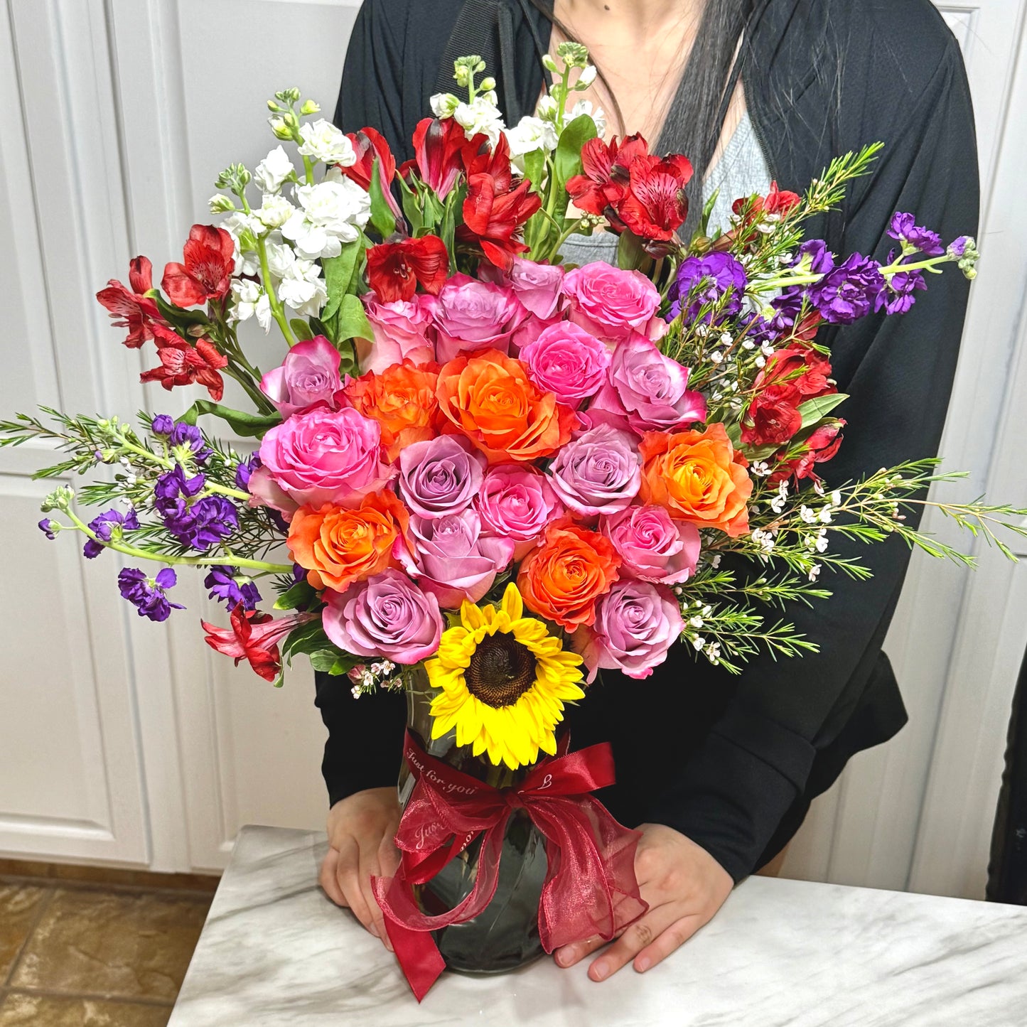 Vibrant bouquet of mixed roses, stock, and a sunflower in a ribbon-tied vase.