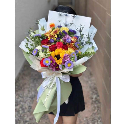 Vibrant bouquet with red roses, yellow sunflowers, and purple flowers wrapped in green paper.