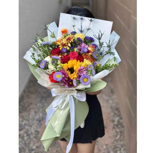 Vibrant bouquet with red roses, yellow sunflowers, and purple flowers wrapped in green paper.