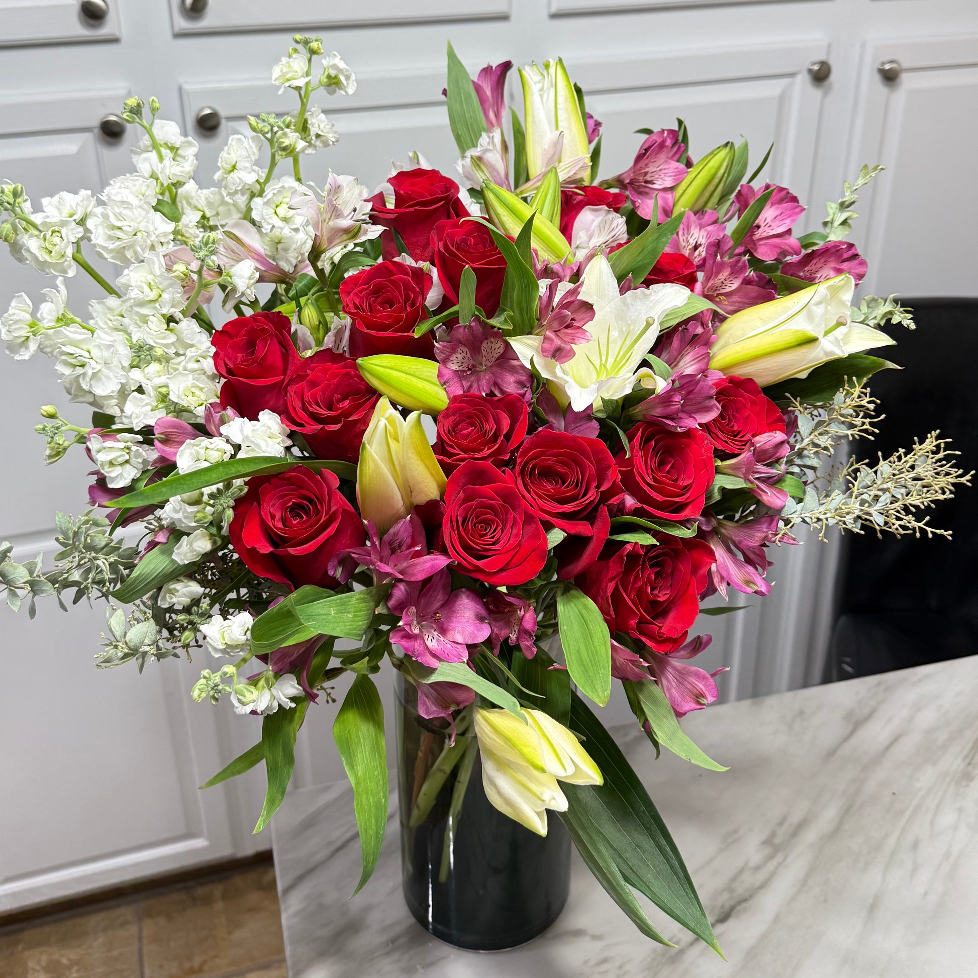 Vibrant bouquet of red roses, white lilies, and stock flowers in a dark vase.