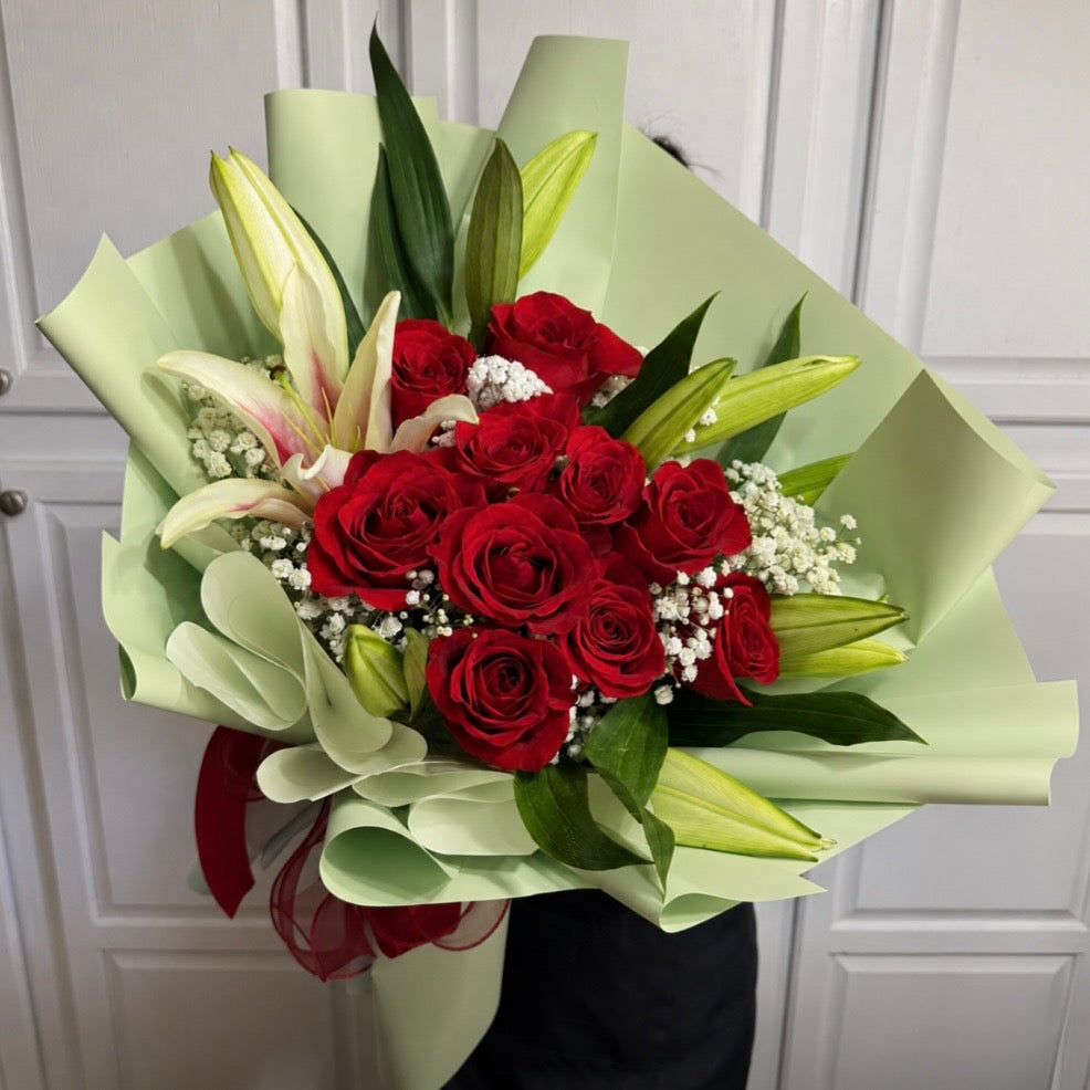 A vibrant bouquet of red roses, white lilies, and baby's breath wrapped in light green paper.