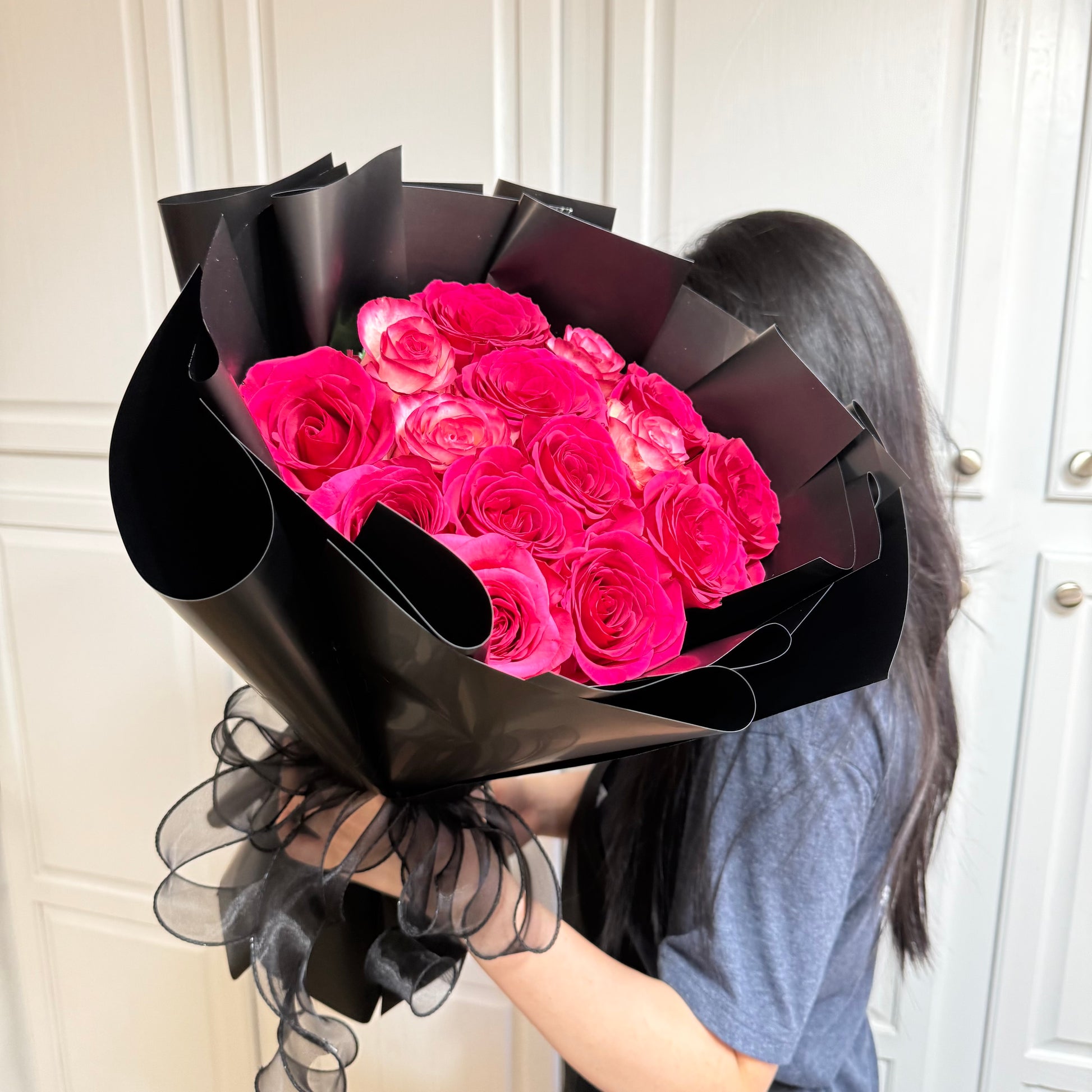 A person holds a bouquet of bright pink roses wrapped in black paper.