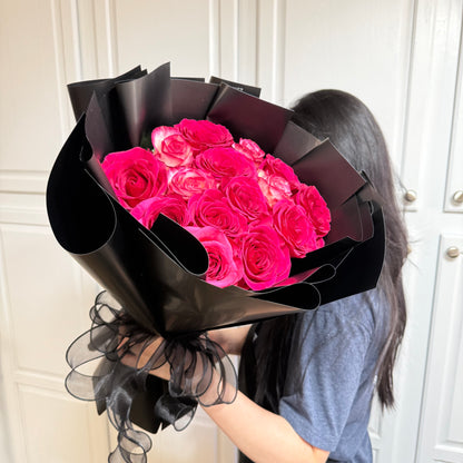 A person holds a bouquet of bright pink roses wrapped in black paper.