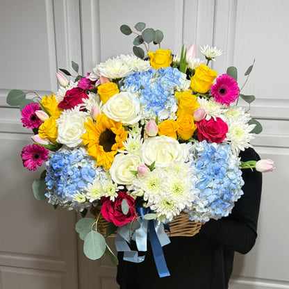 A large, vibrant flower basket with yellow roses, blue hydrangeas, and pink gerberas.