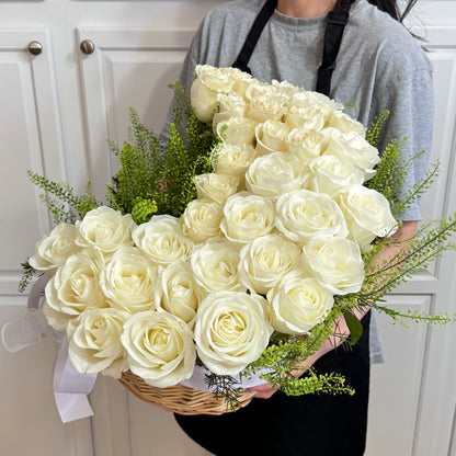 Elegant arrangement of abundant white roses and green foliage in a woven basket.