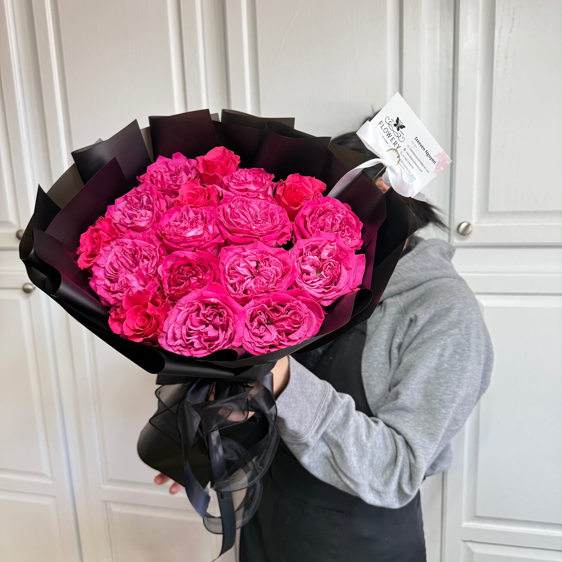 Vibrant pink rose bouquet wrapped in black paper and ribbon, held by a person.