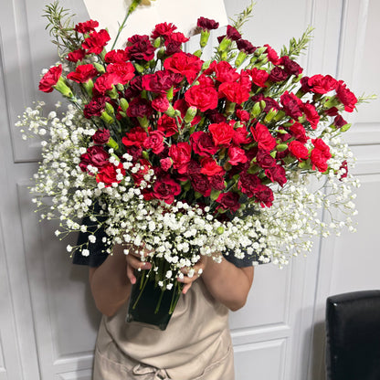 Vibrant red and dark red carnations with delicate white baby's breath in a clear vase.